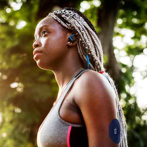 A close up profile shot of a black woman with braids wearing a grey tank top. She's wearing a CGM and a Signos sports cover on the back of her left arm.