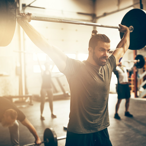 A brown-haired man with a beard is wearing a grey shirt and black shorts and a CGM with a Signos sports cover on his arm. He's lifting a barbell over his head in a gym.