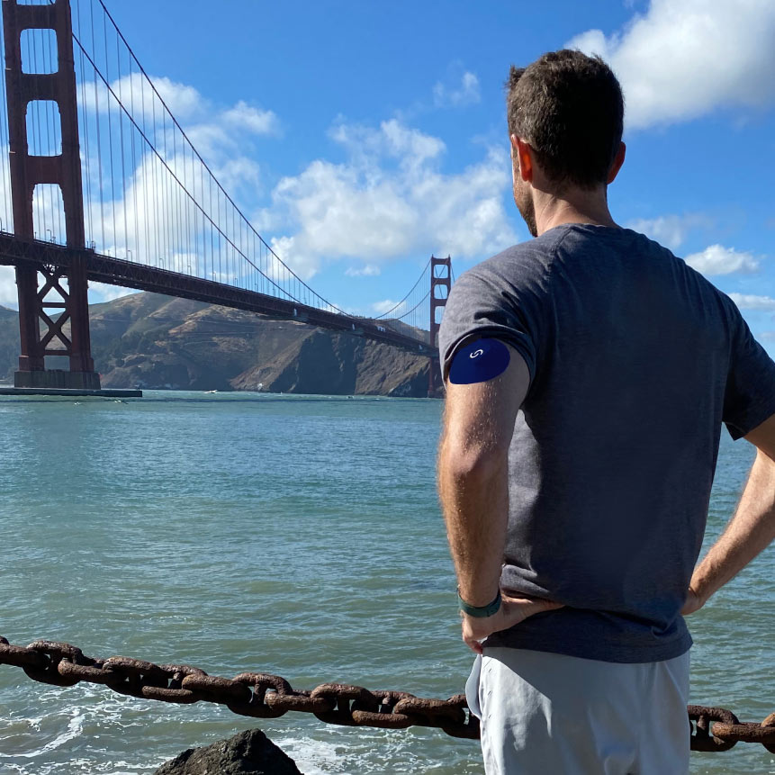 A white man wearing a continuous glucose monitor with a blue Signos sport cover on top on his left arm. His back is facing the camera and he's look at the Golden Gate Bridge.