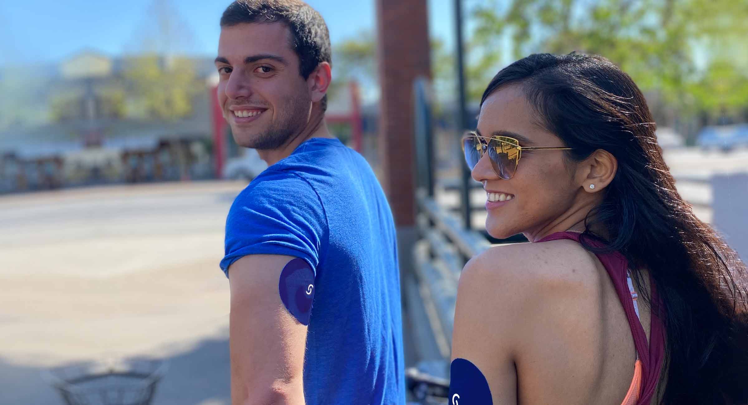 A young Lebanese man and young Indian girl are smiling and looking over their shoulders. Both are wearing a CGM with Signos sports cover on the triceps of their left arms.