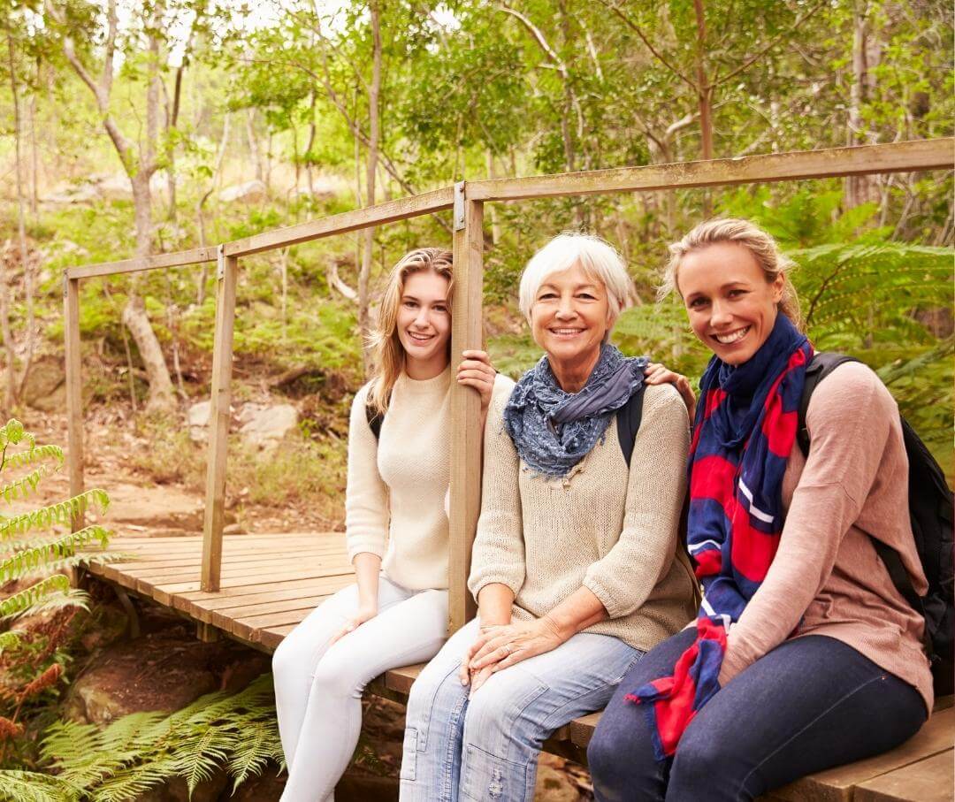 three generations of women sitting on a wooden pedestrian bridge in the forest