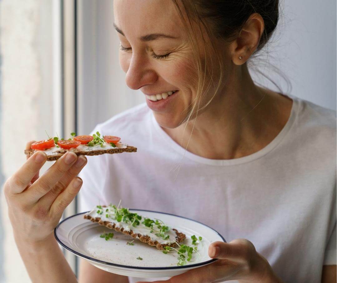 a woman eating a flatbread cracker; topped with tomato, microgreen, and cream cheese