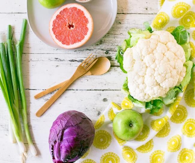 Food on a white table including grapefruit, scallions, purple cabbage, a green apple, and a head of cauliflower