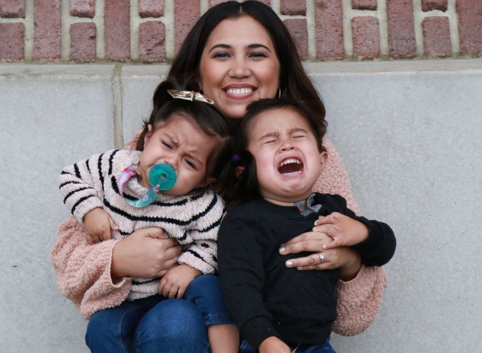 Woman smiling and holding her two children, who are crying