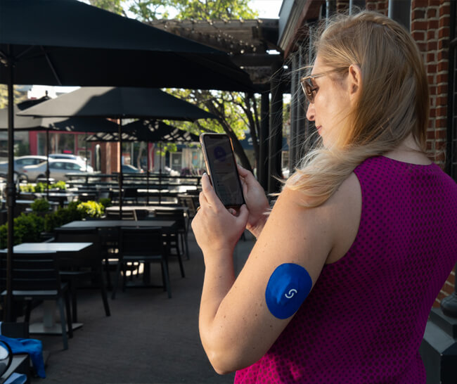 Woman standing outside of a restaurant and pre-tracking her meal in the Signos app. On the back of her left arm is the Signos CGM.