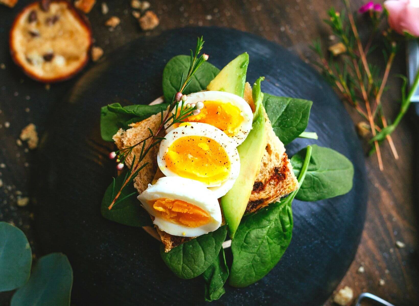 An overhead shot of a black plate with baby spinach, sprouted whole grain toast, sliced avocado, and sliced hardboiled eggs