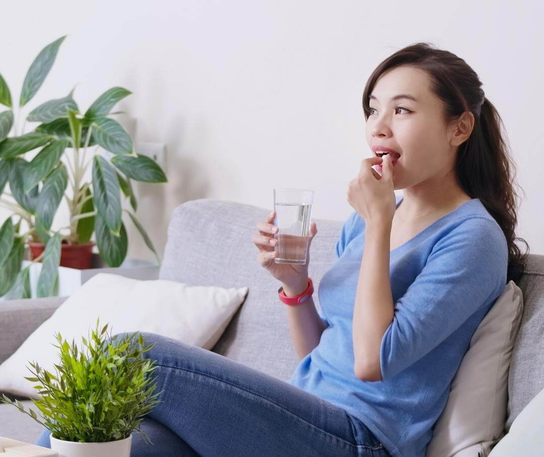 woman sitting on a sofa taking a probiotic supplement with a glass of water