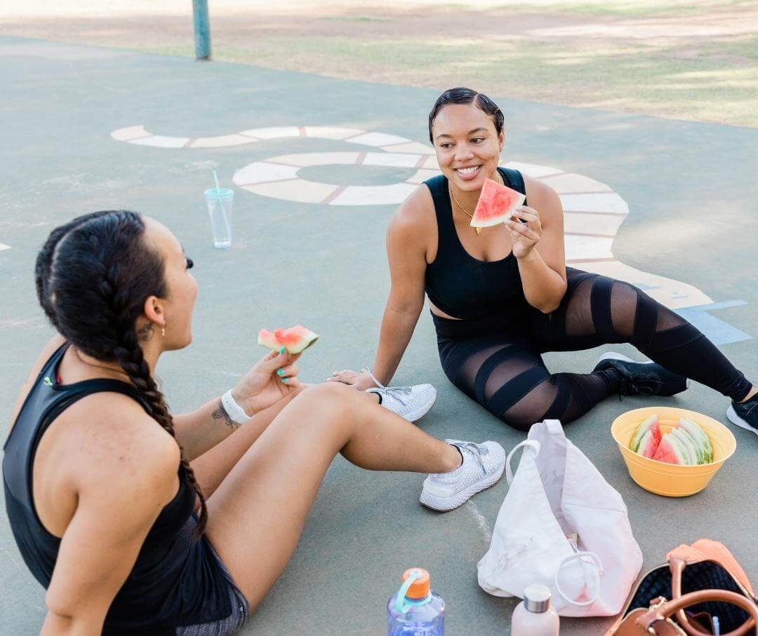 two women sitting on a playground eating watermelon after working out