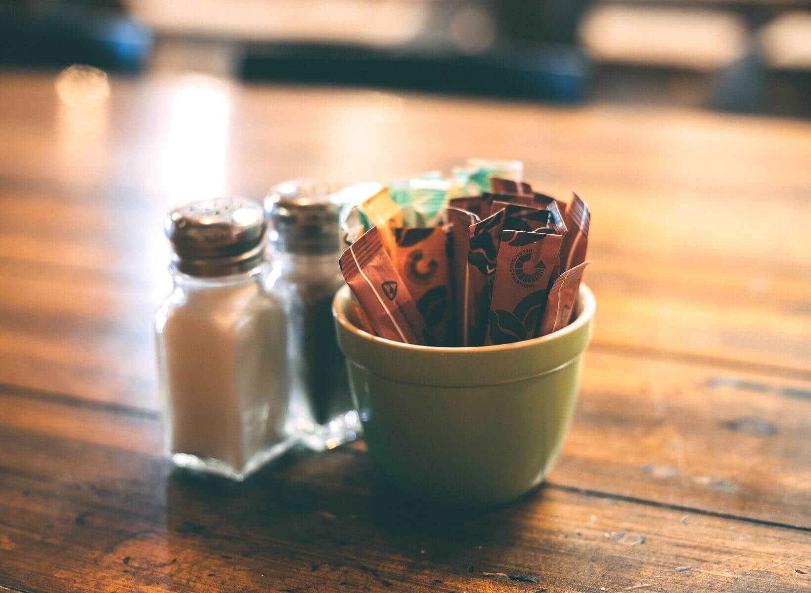 A white ramekin containing sugar and sweeteners like stevia on a wood table next to salt and pepper shakers