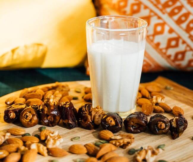 A glass of milk on a wooden cheese board surrounded by nuts and figs