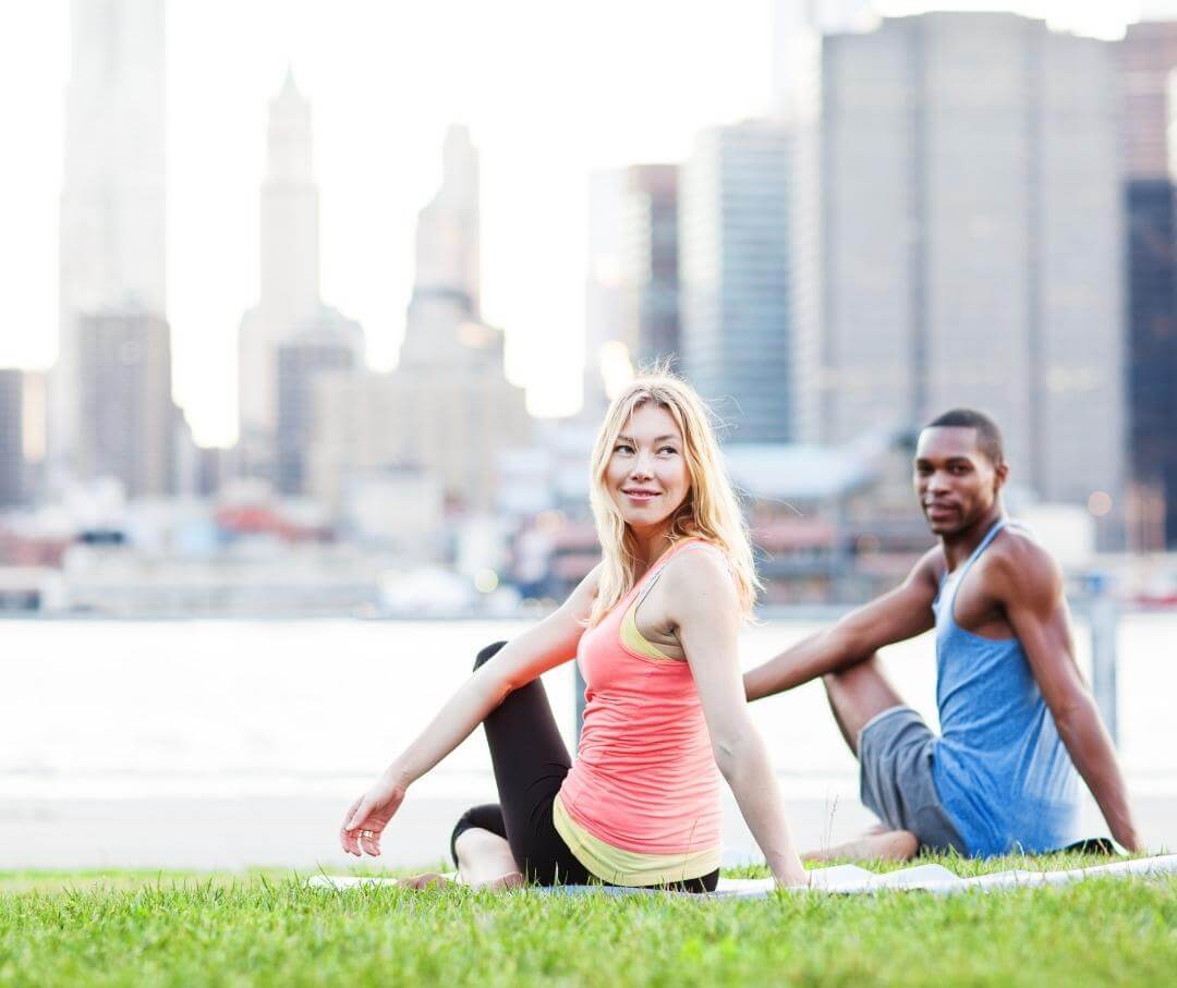 a man and a woman stretching in a grassy area with a cityscape behind them