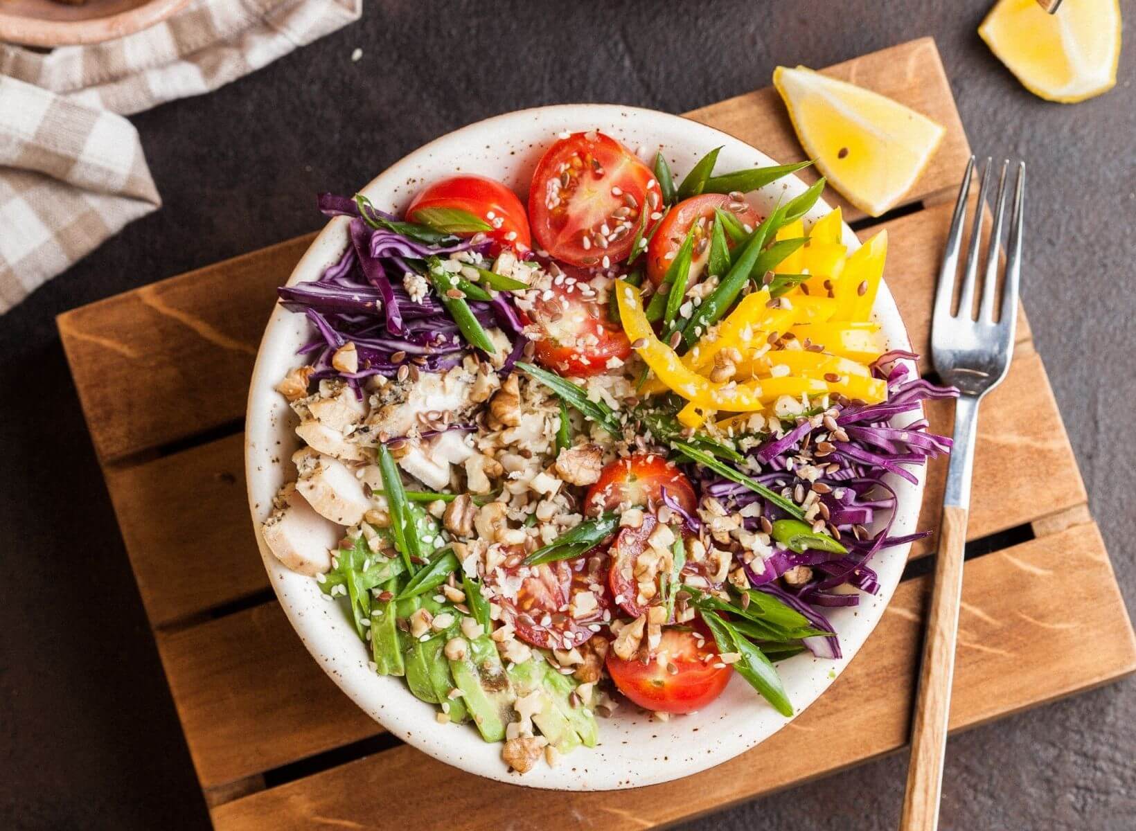 A white bowl filled with low-glycemic vegetables is sitting on a wood cutting board next to a fork and slices of lemon