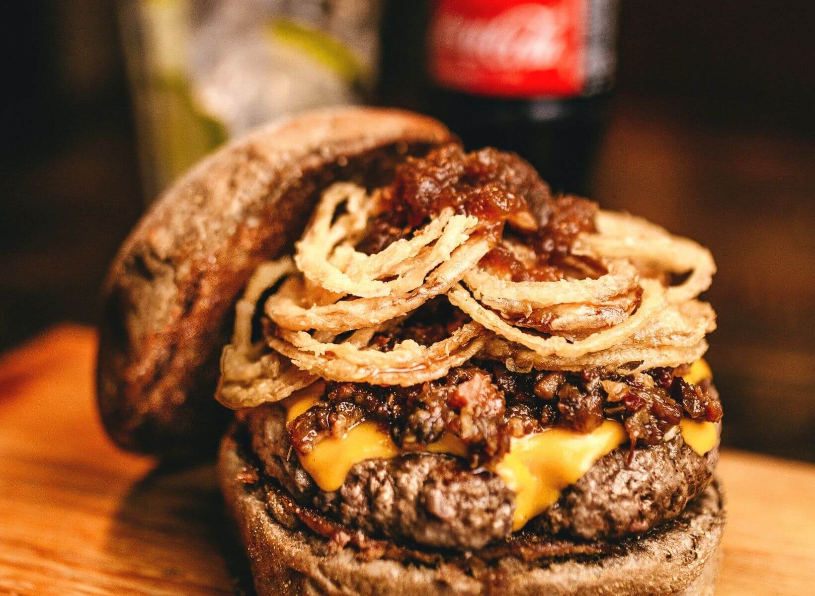 A close up shot of a burger with fried onion rings and a bottle of coke in the background. This food and drink can add to lower belly fat.