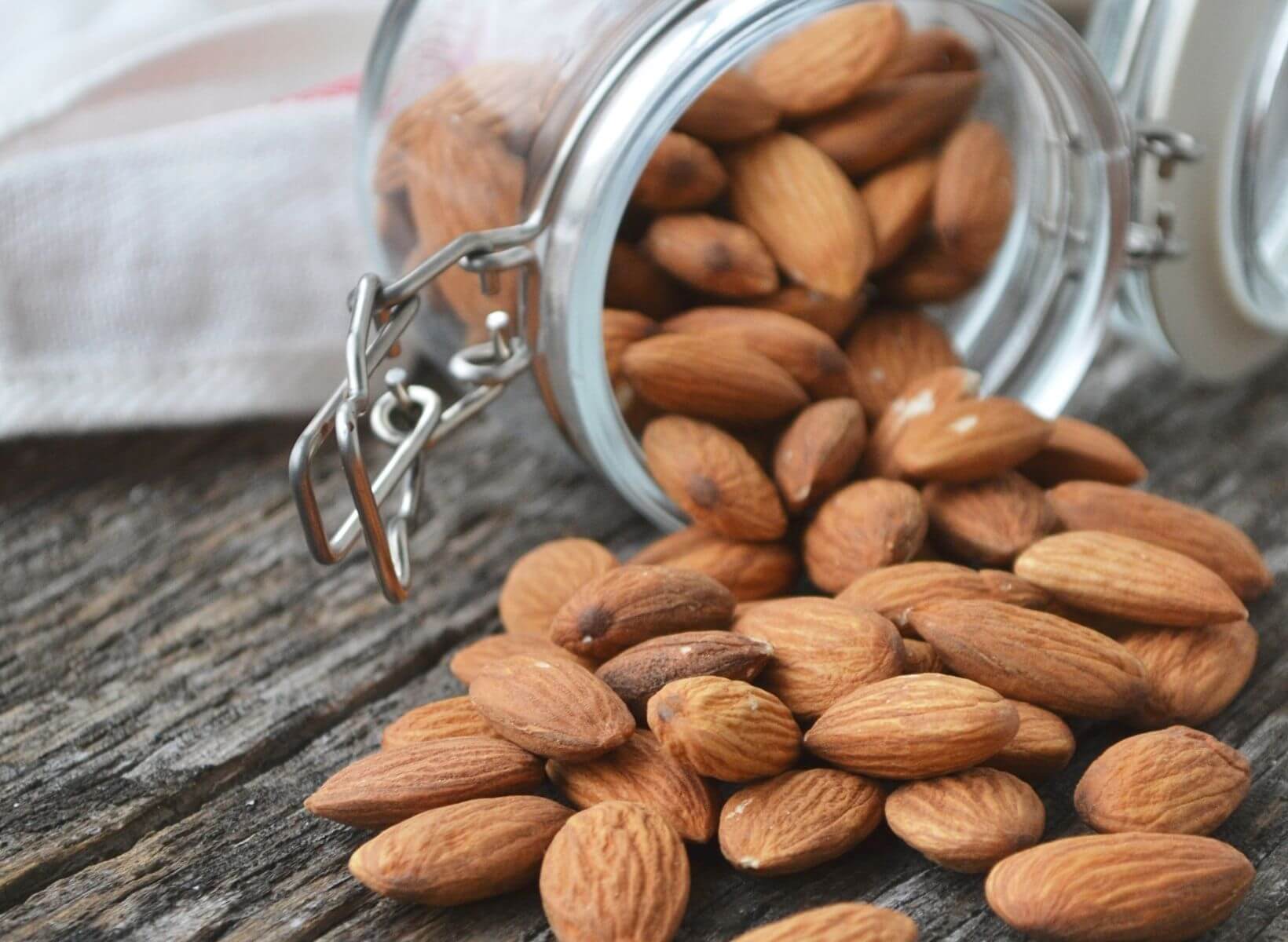 A plastic container with a metal clasp open on its side with raw almonds spilling out. Almonds are a nut source of prebiotics.