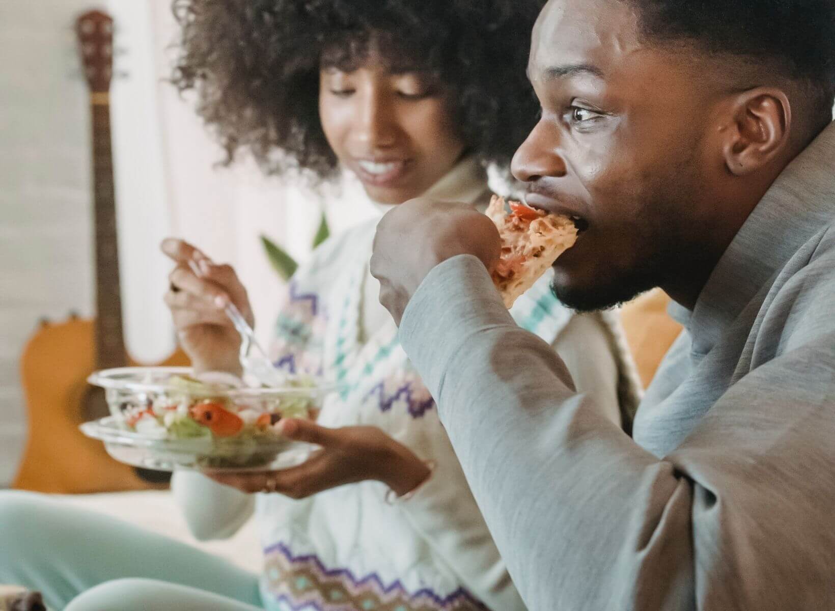 A couple eating dinner on the couch; the woman is eating salad and the man is eating pizza. Photo by Andres Ayrton from Pexels