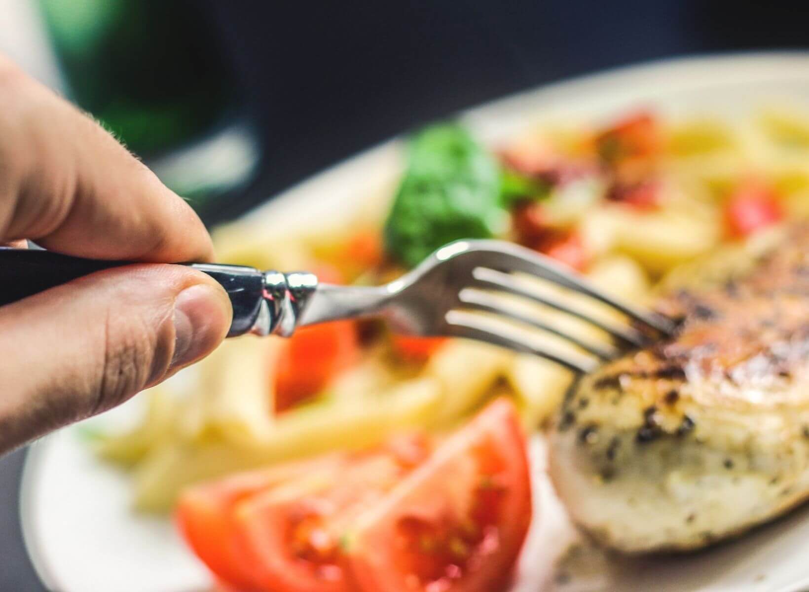 A hand holding a fork poking a cooked chicken breast on a white plate with a tomato slice and blurred vegetables