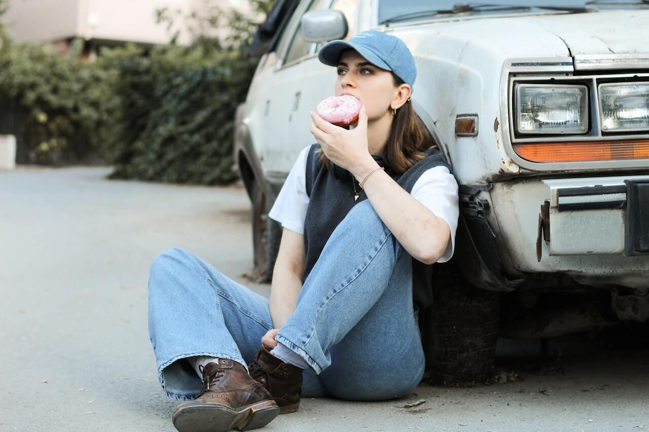 woman-in-cap-sitting-by-car-and-eating-donut