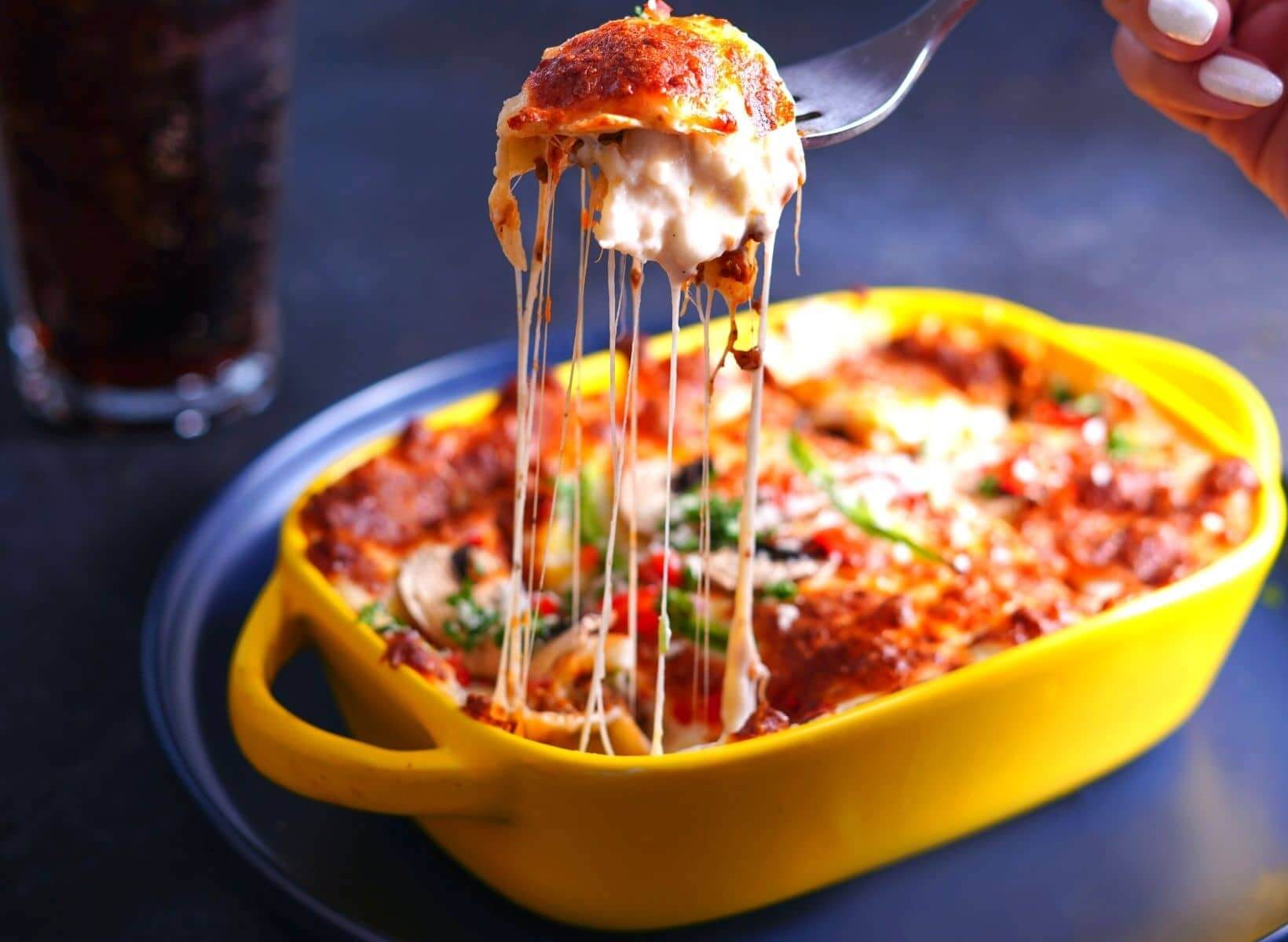 A casserole dish filled with cooked lasagna with a hand pulling a forkful of lasagna with strings of melted cheese hanging down