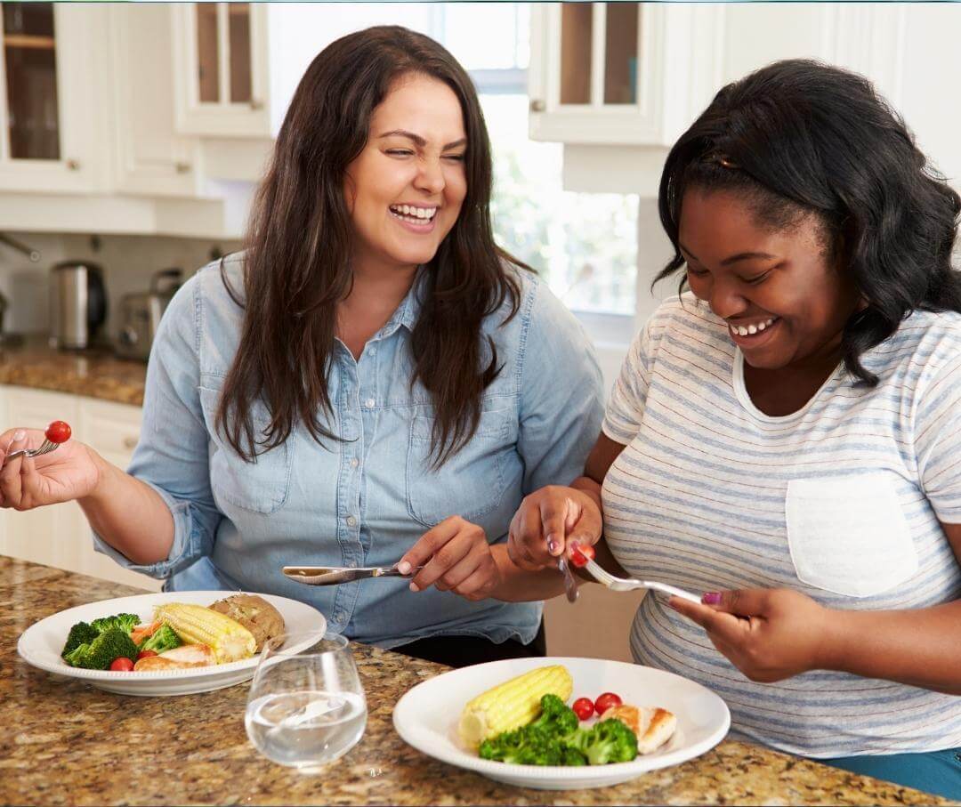 Two women laughing while eating a clean lunch at a kitchen island.