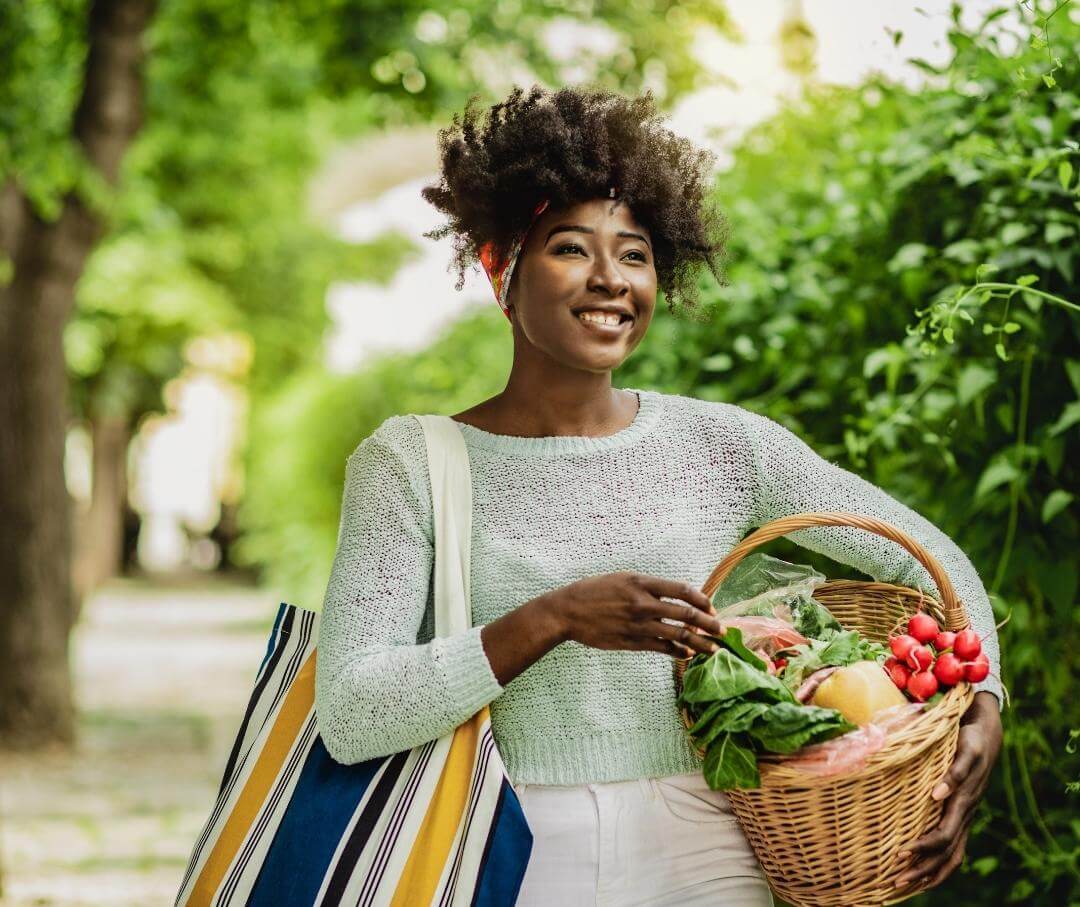 woman walking down a suburban sidewalk with a tote bag and basket of produce