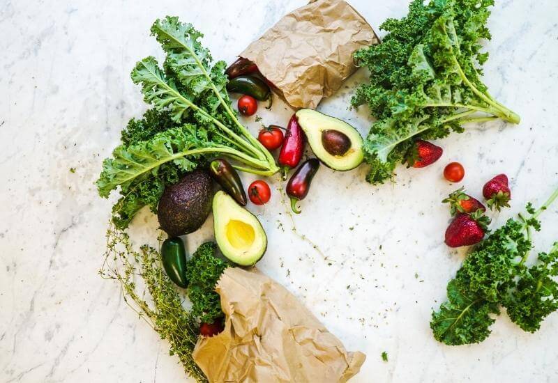 Mixed selection of fruits and vegetables on a marble tabletop, including: avocado, kale, tomatoes