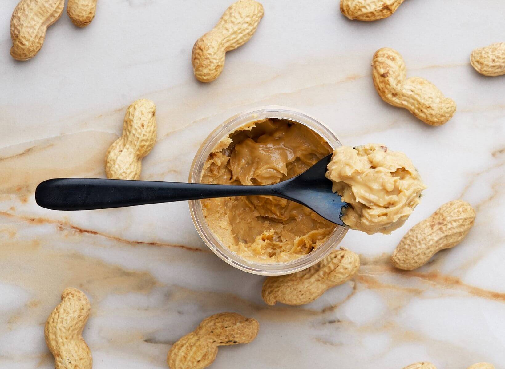 An overhead shot of peanuts in a shell and a jar of crunchy peanut butter with a spoon resting on top. Peanuts and peanut butter are low-glycemic snacks