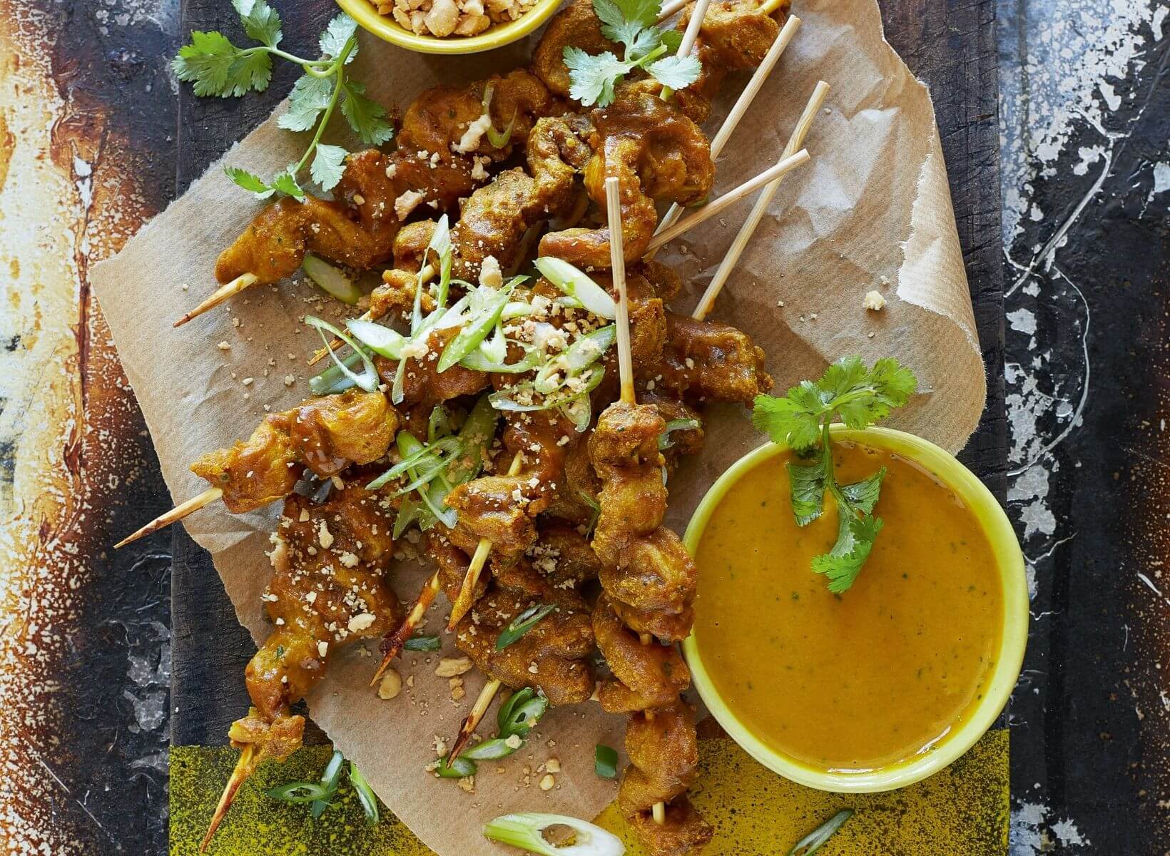 An overhead shot of a board with brown parchment and skewered grilled chicken, an example of a low-glycemic snack. On the side is a ramekin of peanut satay sauce