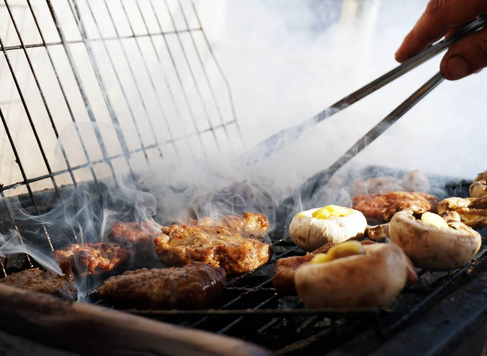 Close up shot of a hand holding tongs, about the flip meat and vegetables on a smoking grill