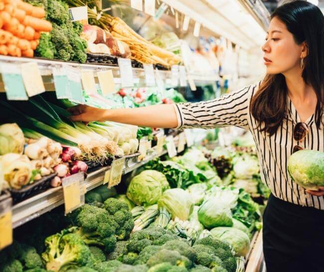 Woman at grocery store holding a head of cabbage and reaching for a leek