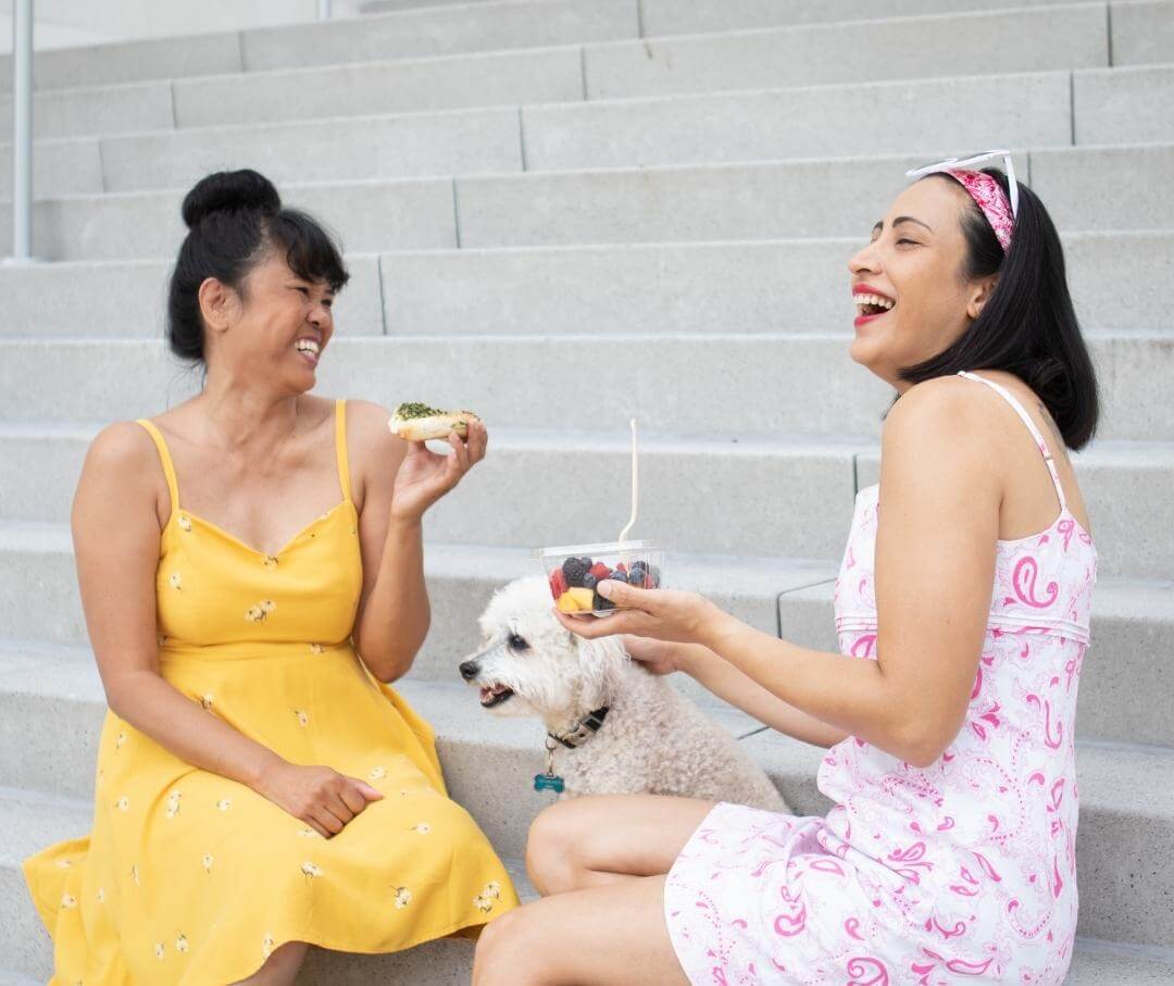 women sitting on some steps outdoors with a small white dog, eating food and laughing